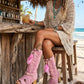 A model wearing the Rhapsody Bohemia Pink Boots at a rustic beach bar. The vibrant pink suede and bohemian details stand out against the sun-drenched wooden deck and coastal decor, embodying a free-spirited summer style.