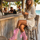 A model wearing the Rhapsody Bohemia Pink Boots at a rustic beach bar. The vibrant pink suede and bohemian details stand out against the sun-drenched wooden deck and coastal decor, embodying a free-spirited summer style.