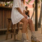 Model at a beach club wearing Atenea beige suede platform boots, showing the natural earthy tones and bohemian details in a sophisticated summer sunset setting