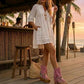 Model wearing Atenea Pink suede platform boots at a beach chiringuito, showcasing the bohemian fringe and shell details in a coastal summer setting.