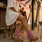 Model wearing Atenea Pink suede platform boots at a beach chiringuito, showcasing the bohemian fringe and shell details in a coastal summer setting.