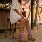 Model wearing Atenea Pink suede platform boots at a beach chiringuito, showcasing the bohemian fringe and shell details in a coastal summer setting.