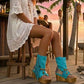 Boho-style model at a beach chiringuito wearing turquoise suede platform flip-flop boots, perfectly complementing a summer beach club outfit.