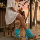 Boho-style model at a beach chiringuito wearing turquoise suede platform flip-flop boots, perfectly complementing a summer beach club outfit.