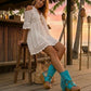 Boho-style model at a beach chiringuito wearing turquoise suede platform flip-flop boots, perfectly complementing a summer beach club outfit.