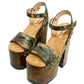 Side view of Crazy Armi Sandals by Sol Caleyo, featuring a 17 cm wooden heel, camouflage leather straps, and brass stud details on a white background.