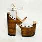 Side view of Crazy White Sandals by Sol Caleyo, featuring white leather straps with bronze studs on a high dark wood platform and tractor sole.