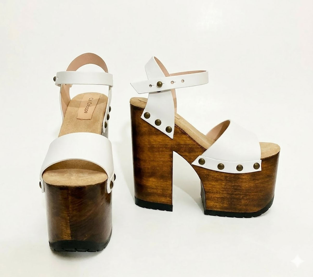 Side view of Crazy White Sandals by Sol Caleyo, featuring white leather straps with bronze studs on a high dark wood platform and tractor sole.