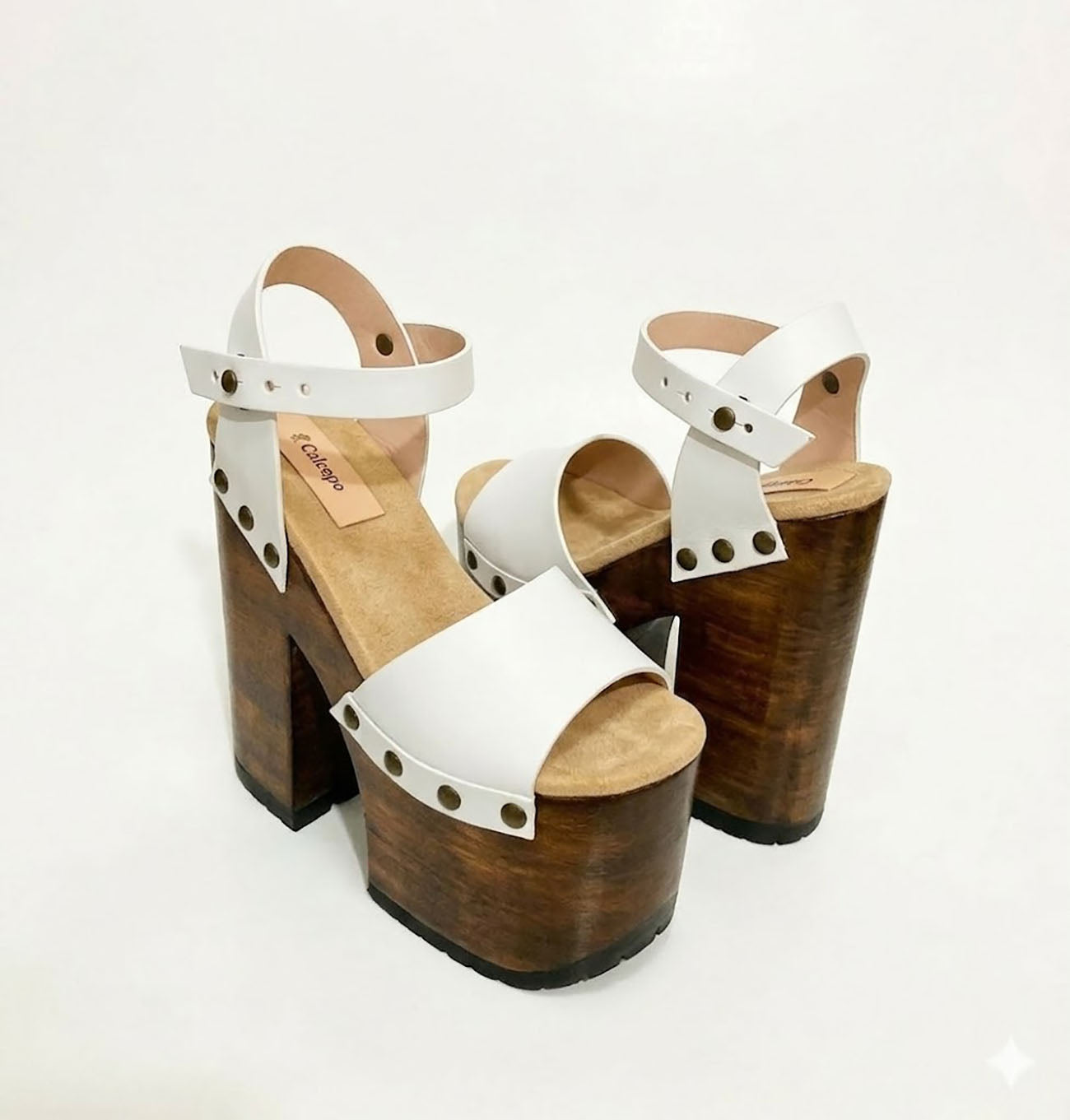Side view of Crazy White Sandals by Sol Caleyo, featuring white leather straps with bronze studs on a high dark wood platform and tractor sole.