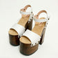 Side view of Crazy White Sandals by Sol Caleyo, featuring white leather straps with bronze studs on a high dark wood platform and tractor sole.