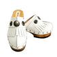 Top view of a pair of handmade white textured full-grain leather clogs with leather fringes and detailed Native American chief profile metal conchos on a wooden platform.