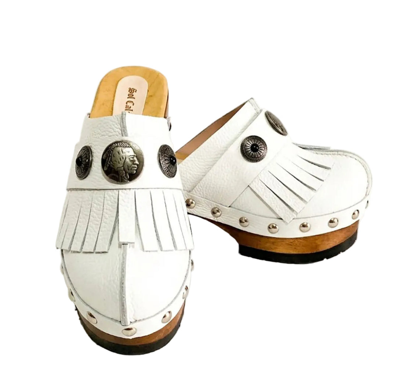 Top view of a pair of handmade white textured full-grain leather clogs with leather fringes and detailed Native American chief profile metal conchos on a wooden platform.