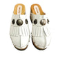Top view of a pair of handmade white textured full-grain leather clogs with leather fringes and detailed Native American chief profile metal conchos on a wooden platform.
