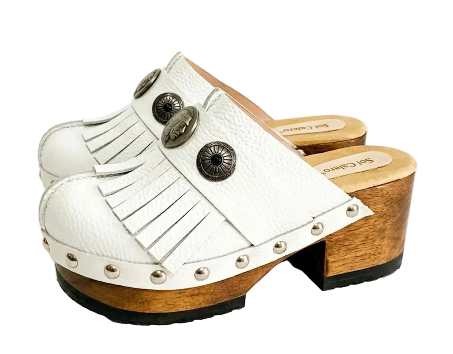 Top view of a pair of handmade white textured full-grain leather clogs with leather fringes and detailed Native American chief profile metal conchos on a wooden platform.