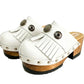 Top view of a pair of handmade white textured full-grain leather clogs with leather fringes and detailed Native American chief profile metal conchos on a wooden platform.