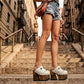 Close-up of a model wearing Sol Caleyo White Indian Clogs XL with 17cm heels and metal conchos, styled with denim shorts in an urban setting.