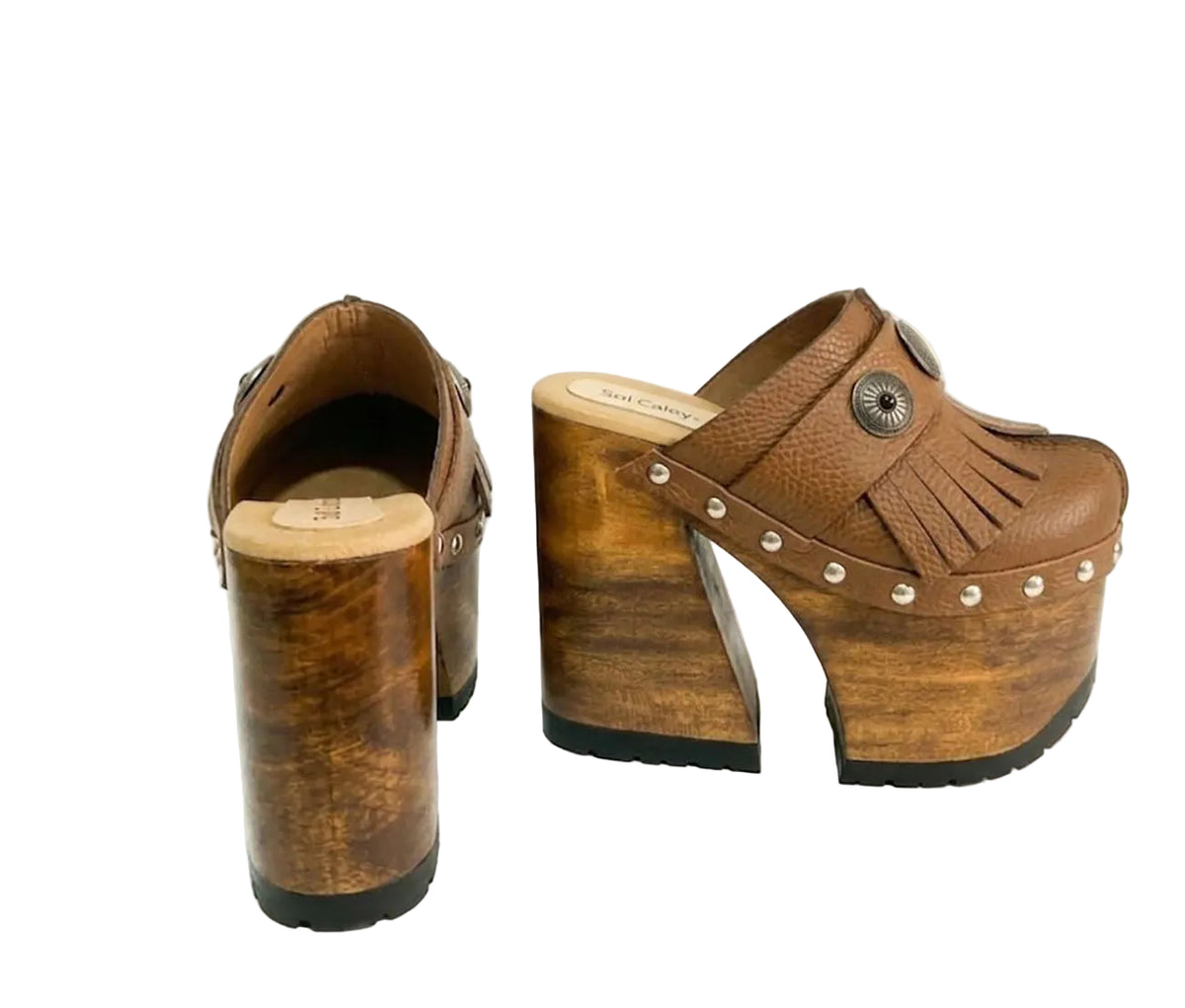 Side profile of Sol Caleyo Light Brown Indian Clogs XL featuring a high 17cm wooden platform, textured brown leather, decorative fringes, and antique metal conchos on a white background.