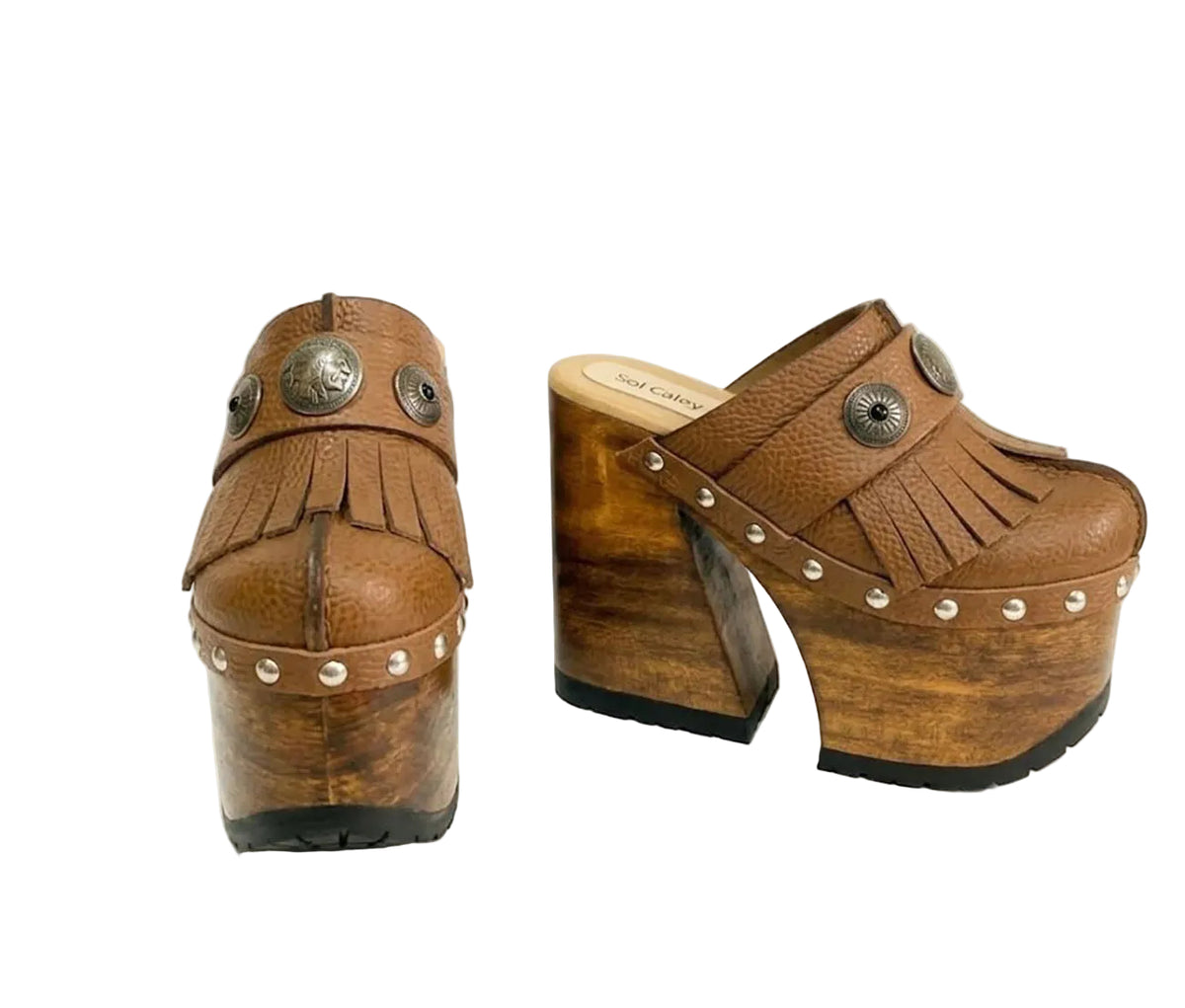Side profile of Sol Caleyo Light Brown Indian Clogs XL featuring a high 17cm wooden platform, textured brown leather, decorative fringes, and antique metal conchos on a white background.
