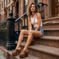 Fashion model sitting on urban brownstone steps wearing Light Brown Indian Clogs XL with denim shorts and a white tank top, showcasing the extreme wooden platform height.