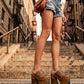Fashion model sitting on urban brownstone steps wearing Light Brown Indian Clogs XL with denim shorts and a white tank top, showcasing the extreme wooden platform height.