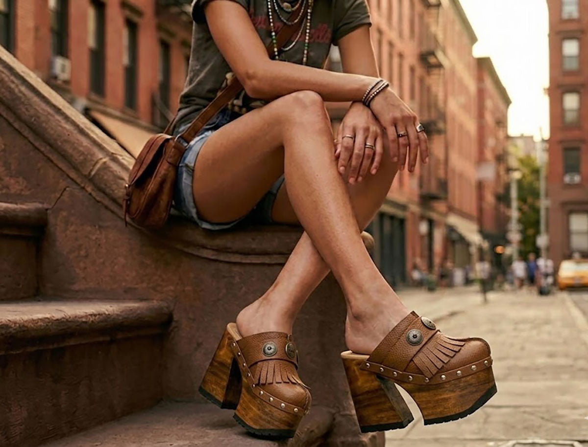 Fashion model sitting on urban brownstone steps wearing Light Brown Indian Clogs XL with denim shorts and a white tank top, showcasing the extreme wooden platform height.