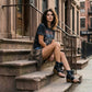 Model sitting on city stairs wearing Sol Caleyo Texas Boots: black leather flip-flop platform sandals with silver studs and chains. Handcrafted 9 cm wooden wedge with a rugged biker aesthetic.