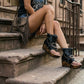 Model sitting on city stairs wearing Sol Caleyo Texas Boots: black leather flip-flop platform sandals with silver studs and chains. Handcrafted 9 cm wooden wedge with a rugged biker aesthetic.