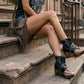 Model sitting on city stairs wearing Sol Caleyo Texas Boots: black leather flip-flop platform sandals with silver studs and chains. Handcrafted 9 cm wooden wedge with a rugged biker aesthetic.