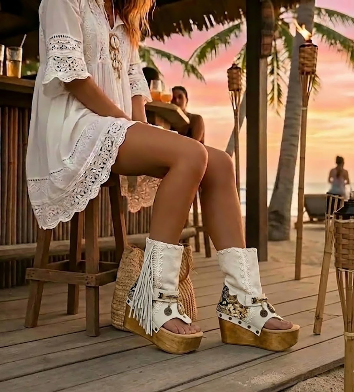 Boho-chic model at a beach club wearing Atenea White suede platform boots, showcasing the white fringe and artisanal details in a sunny, seaside atmosphere.