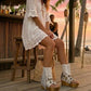 Boho-chic model at a beach club wearing Atenea White suede platform boots, showcasing the white fringe and artisanal details in a sunny, seaside atmosphere.