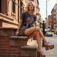 Close-up image of a model from the waist down wearing the Luxury Queen Tattoo Sandals by Sol Caleyo and rolled-up dark denim jeans, posing on a classic brownstone stone staircase in New York City.