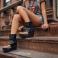 Model wearing Sol Caleyo Texas artisan sandals. High-end platform footwear in black leather with silver hardware and studded details. Luxury handmade wooden wedge sandals for a bold bohemian look.