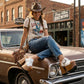 A woman with a cowgirl aesthetic sitting on the hood of a vintage car, wearing premium white leather Western platform clogs by Sol Caleyo with carved wooden soles.