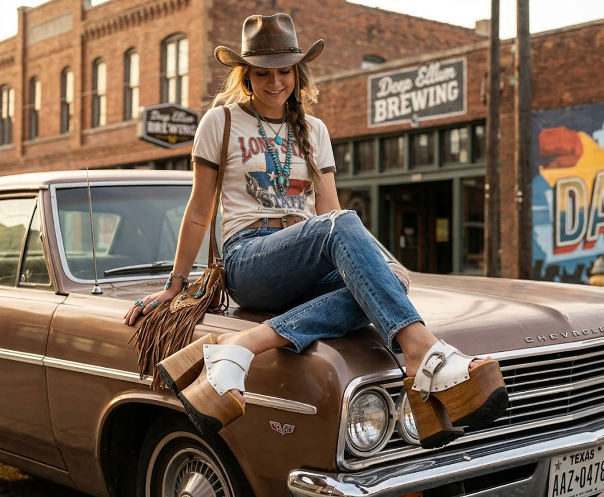 A woman with a cowgirl aesthetic sitting on the hood of a vintage car, wearing premium white leather Western platform clogs by Sol Caleyo with carved wooden soles.