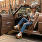 Full-body shot of a woman wearing a cowboy hat and handmade white leather Western platform clogs by Sol Caleyo, posing in a classic car, highlighting nomad-chic aesthetic.