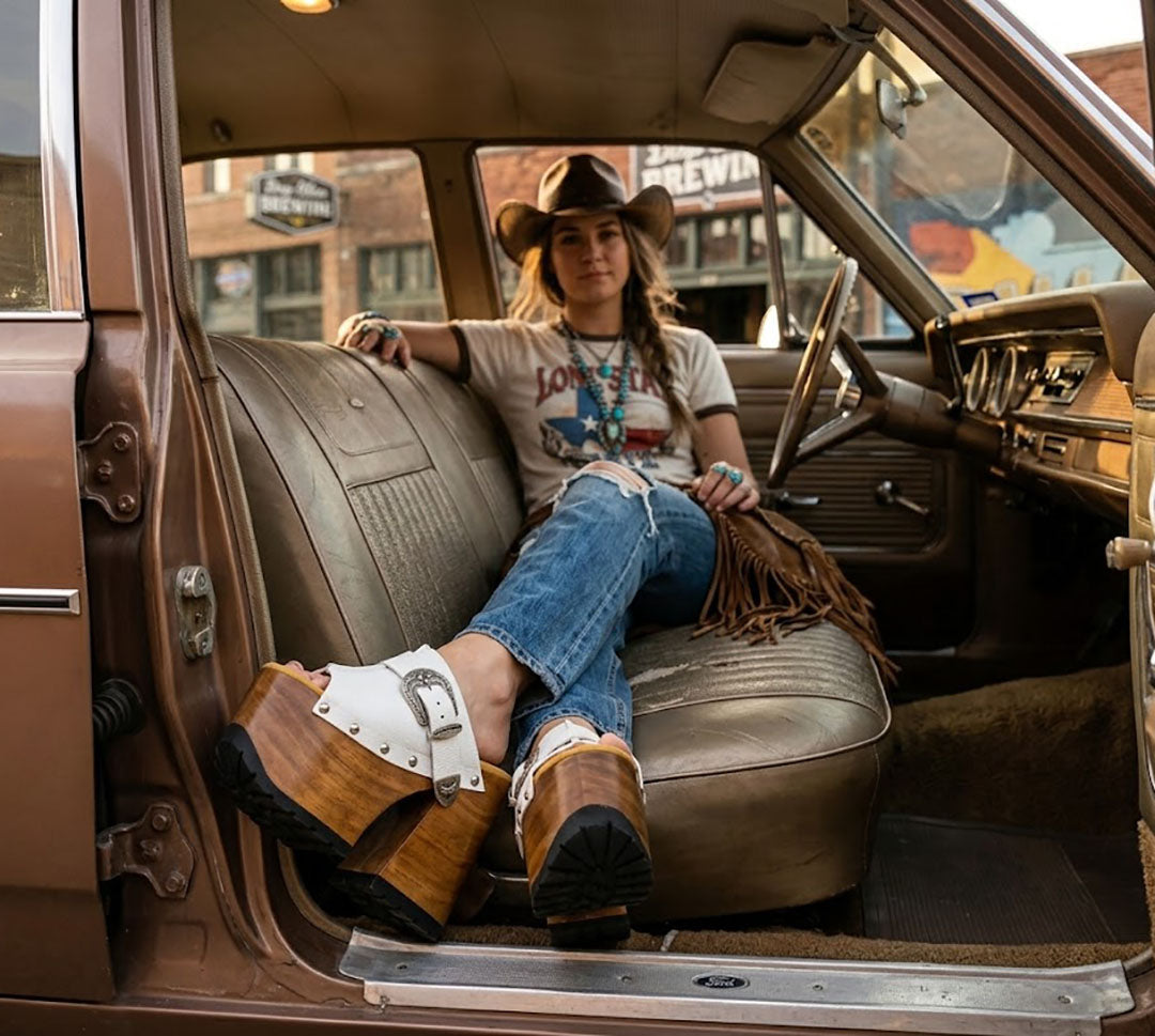 A woman in cowgirl attire wearing handmade white leather Western platform clogs by Sol Caleyo, sitting inside a vintage truck, showcasing artisan wooden heels and boho style.