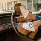 Close-up view of handmade white leather Western platform clogs by Sol Caleyo, featuring an extreme wooden heel, antique brass buckle, and metallic stud details inside a vintage car.