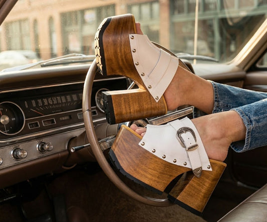 Close-up view of handmade white leather Western platform clogs by Sol Caleyo, featuring an extreme wooden heel, antique brass buckle, and metallic stud details inside a vintage car.