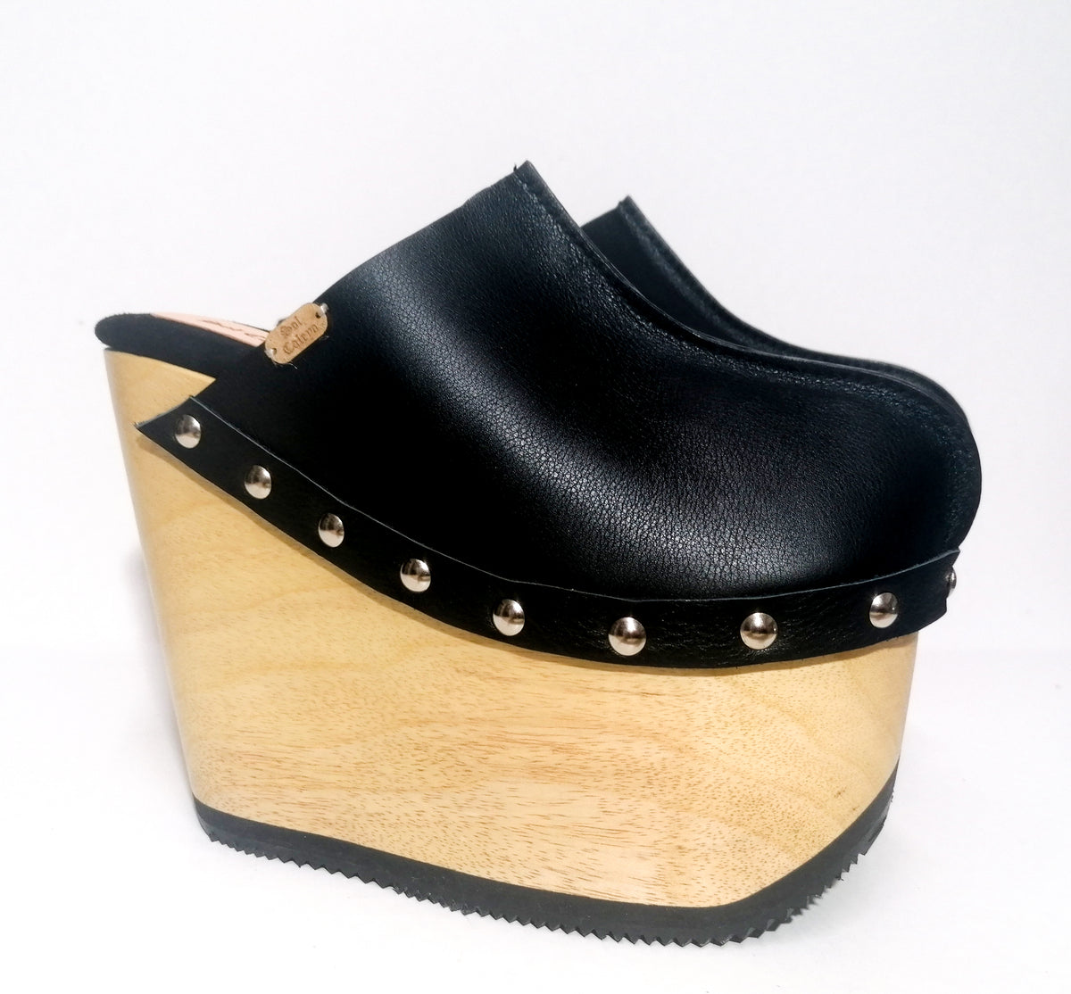 Handmade smooth black leather closed-toe wedge mule clogs with round flat silver studs and 15 cm light natural lacquered wooden wedge by Sol Caleyo.