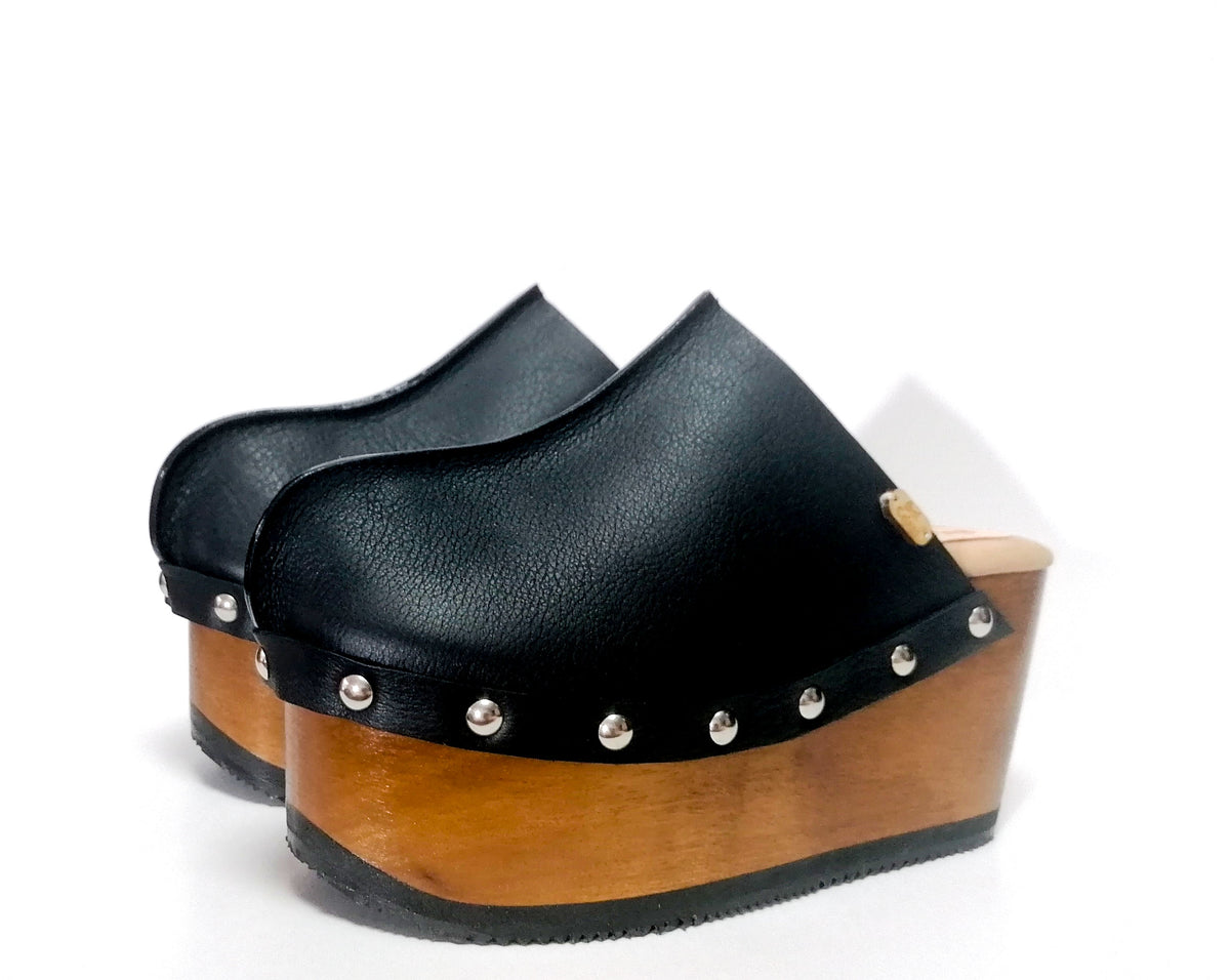 Handmade smooth black leather closed-toe wedge mule clogs with round flat silver studs and 9 cm natural lacquered wooden wedge by Sol Caleyo.