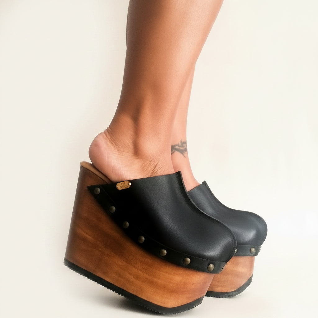 Handmade black leather closed-toe wedge mule clogs with round flat old-gold studs and 15 cm oak-colored lacquered wooden wedge by Sol Caleyo.
